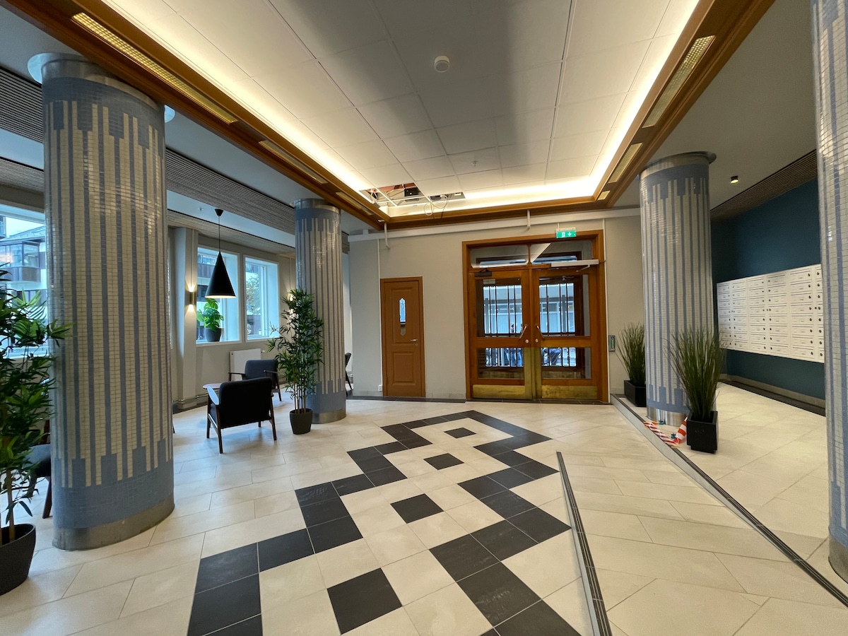A modern lobby area features tall columns with decorative accents. A geometric tile pattern adorns the floor, leading toward the entrance doors. Potted plants are placed strategically, and seating is available. Soft lighting enhances the welcoming ambiance of the space.