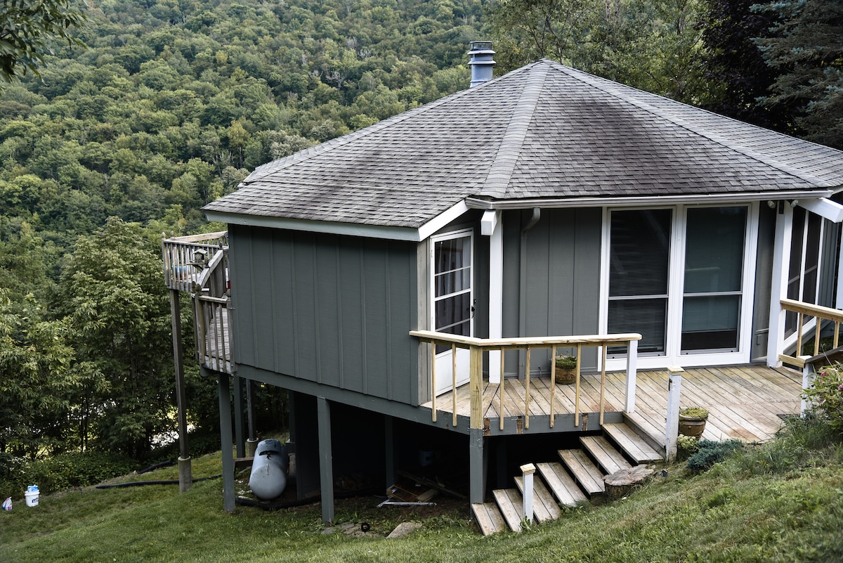 A chalet exterior is showcased, featuring a sloped roof and wooden deck. The structure is surrounded by greenery, providing a sense of seclusion. A set of stairs leads to the entrance, while the deck offers space for outdoor seating.