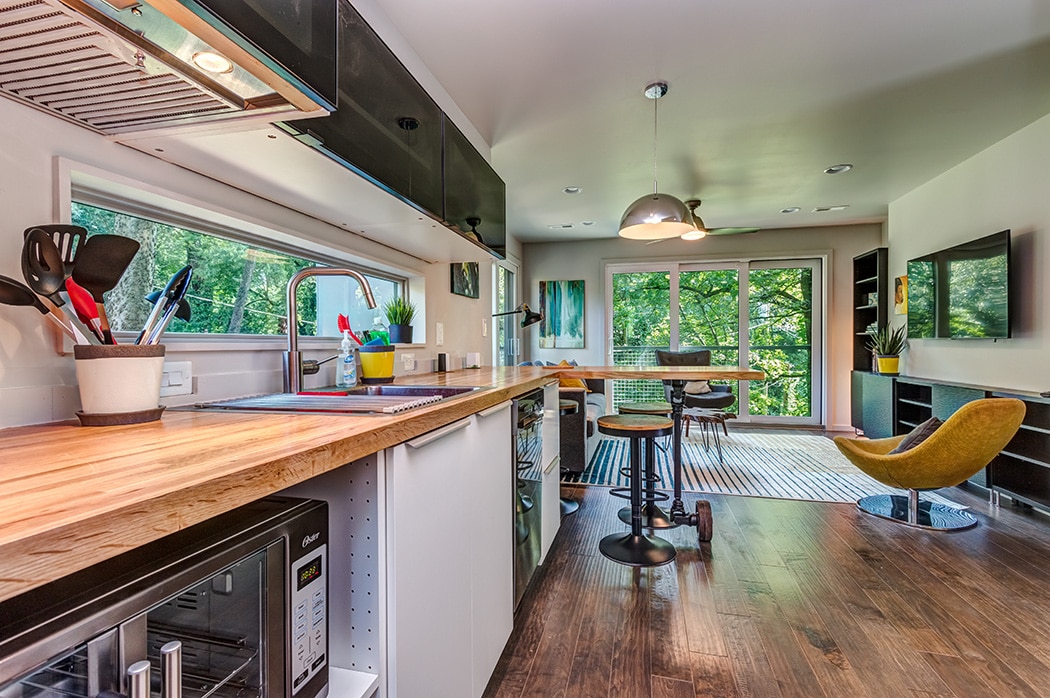 A modern kitchenette with custom butcher block countertops features stainless-steel appliances and a large window providing a view of the lush greenery outside. An island with seating extends into the open living area, highlighted by large glass doors that enhance natural light.