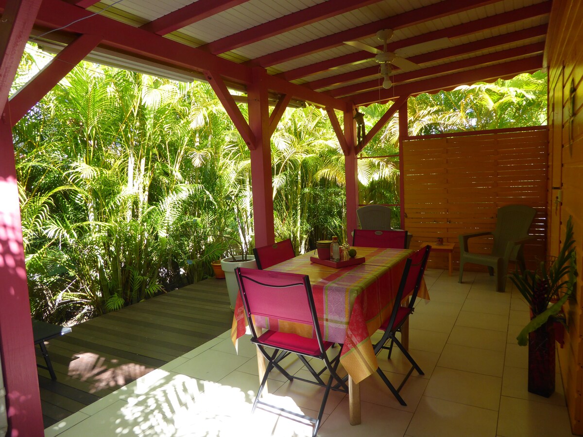 A shaded terrace is surrounded by lush greenery, offering a sense of privacy. A large table, draped with a colorful tablecloth, is flanked by chairs in bright pink. Two additional chairs with a simple design are located nearby, creating a comfortable space for outdoor dining.