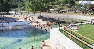 A natural swimming area is displayed, featuring clear water surrounded by a sandy beach and grassy banks. Families and individuals are seen enjoying the space, some relaxing on the grass and others engaging in water activities. Lush trees provide shade in the background.