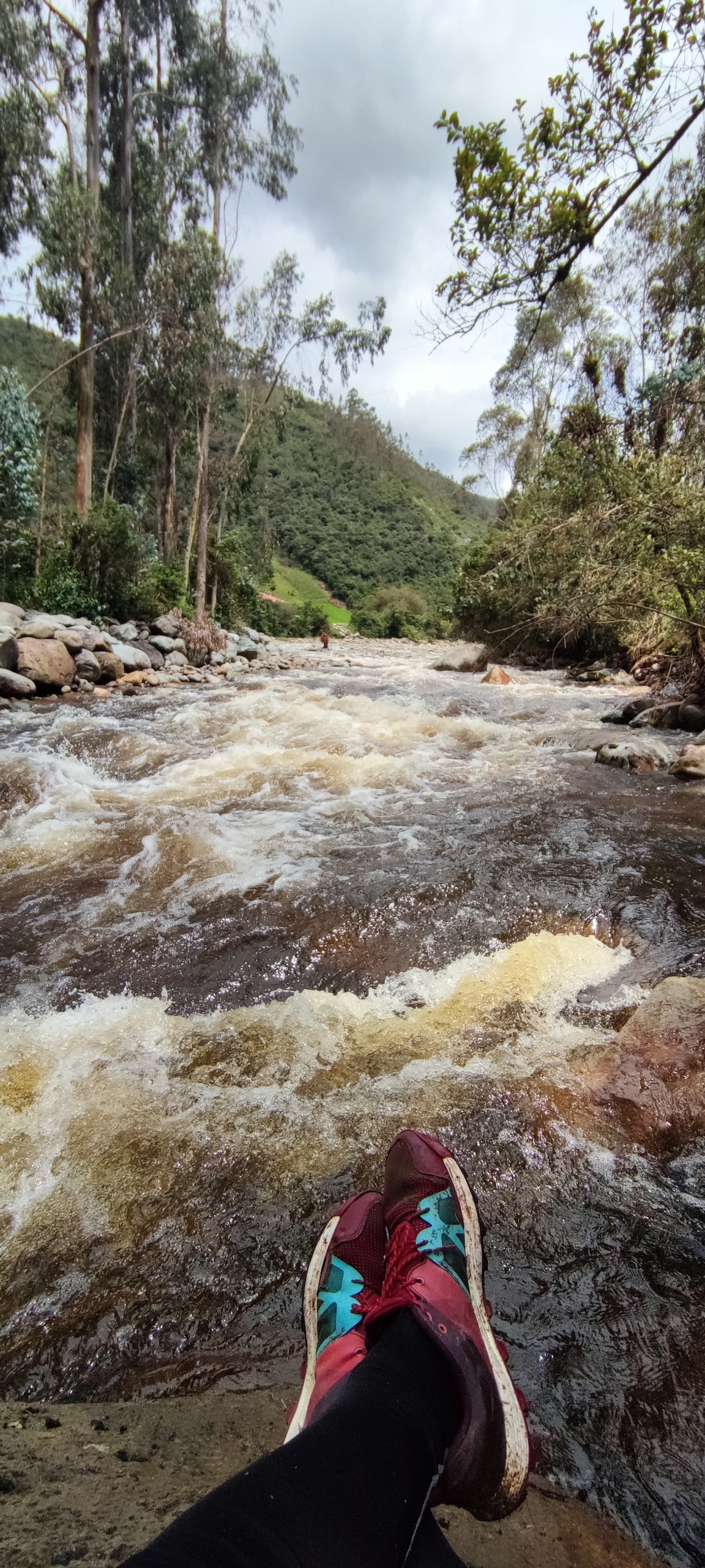 A river flows gently over smooth stones, surrounded by lush greenery and distant hills. The scene captures a pair of colorful shoes resting on the riverbank, with water cascading nearby. The atmosphere reflects a serene natural setting, inviting moments of relaxation and contemplation.