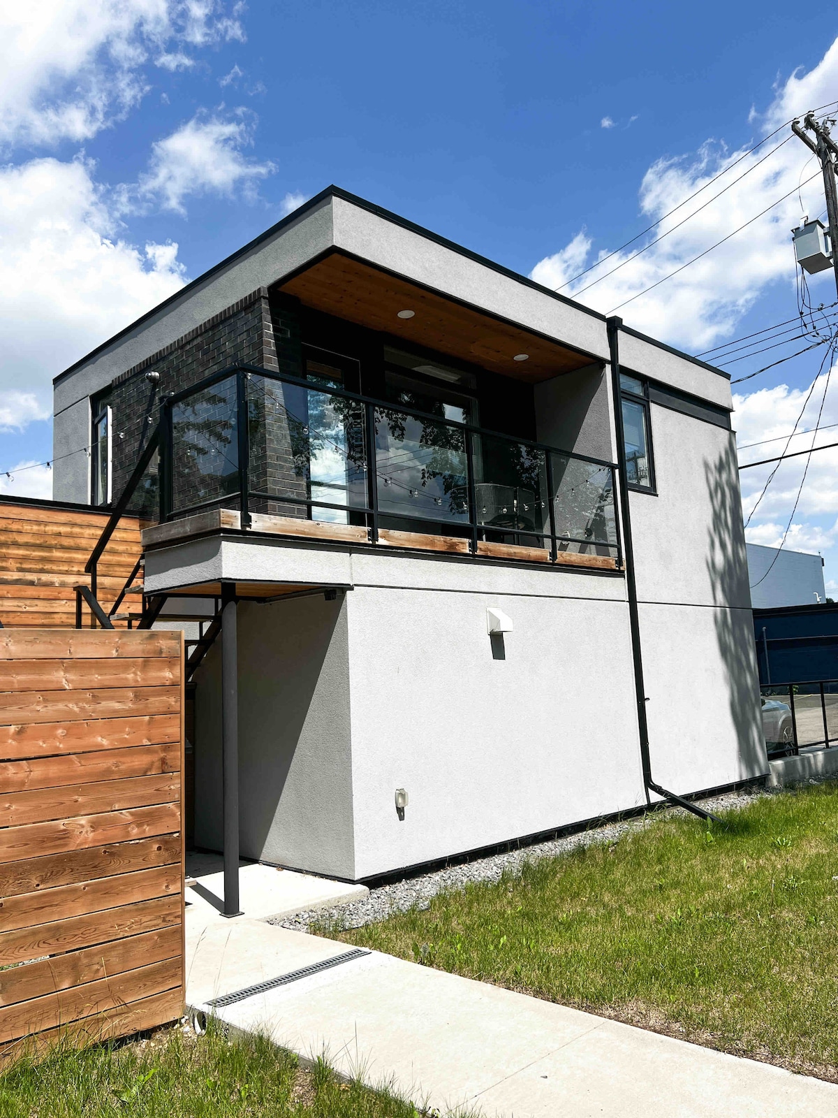 The exterior of the modern building showcases a sleek design with a mix of materials, including smooth stucco and wood accents. A glass balcony extends from the upper level, while a pathway leads to the entrance. Lush green grass surrounds the structure, enhancing its contemporary appeal.