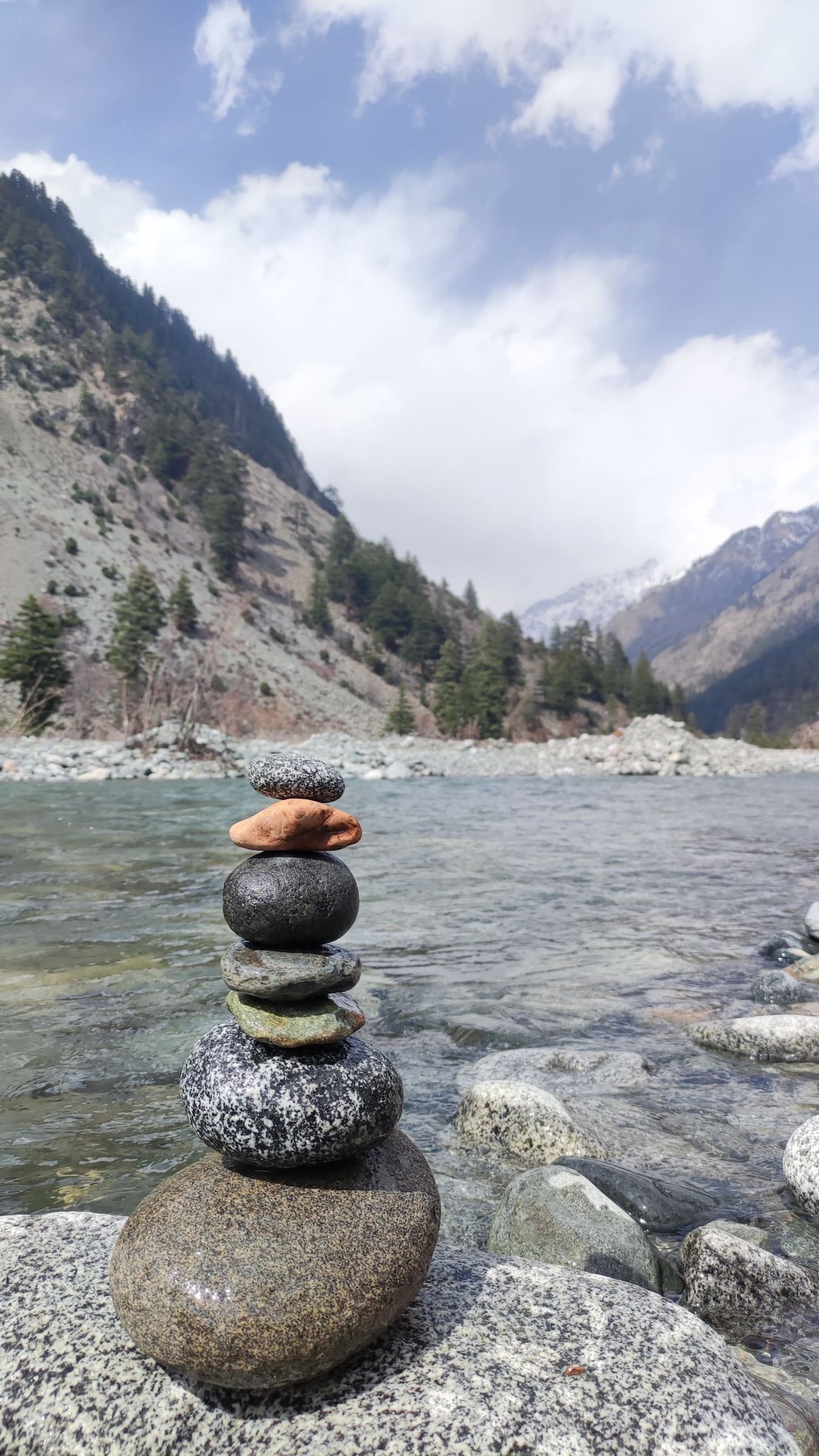 A stack of smooth stones rests on larger rocks by a gently flowing river. Lush greenery lines the riverbanks, leading up to mountains in the background. The sky is partly cloudy, with hints of blue peeking through.