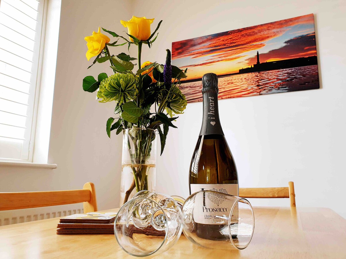 A dining table is set with a bottle of Prosecco and two glasses positioned on a wooden surface. A vase holds fresh yellow roses and greenery. The backdrop displays a vibrant sunset image featuring a lighthouse.