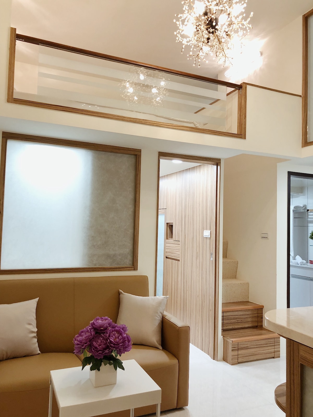 A light and airy living space features a beige sofa adorned with soft pillows. Above, a sparkling chandelier is visible, accompanied by a glass railing that overlooks the upper level. The subtle decor and open layout contribute to a welcoming environment.