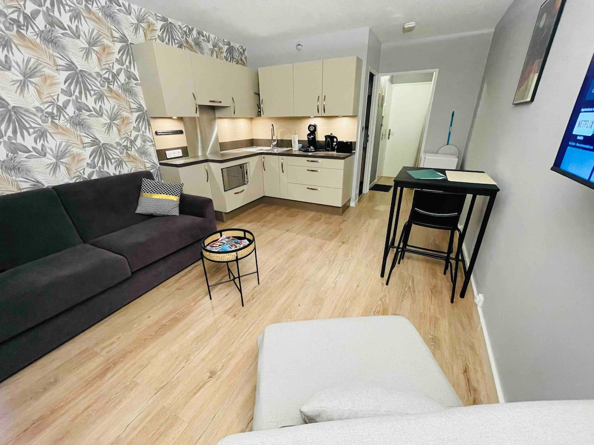A bright studio features a well-equipped kitchen with modern appliances. A dark sofa is positioned opposite a tall dining table with stools. Light wood flooring complements the contemporary decor, which includes a tropical-themed wall. A television is mounted on the wall, enhancing the living area.
