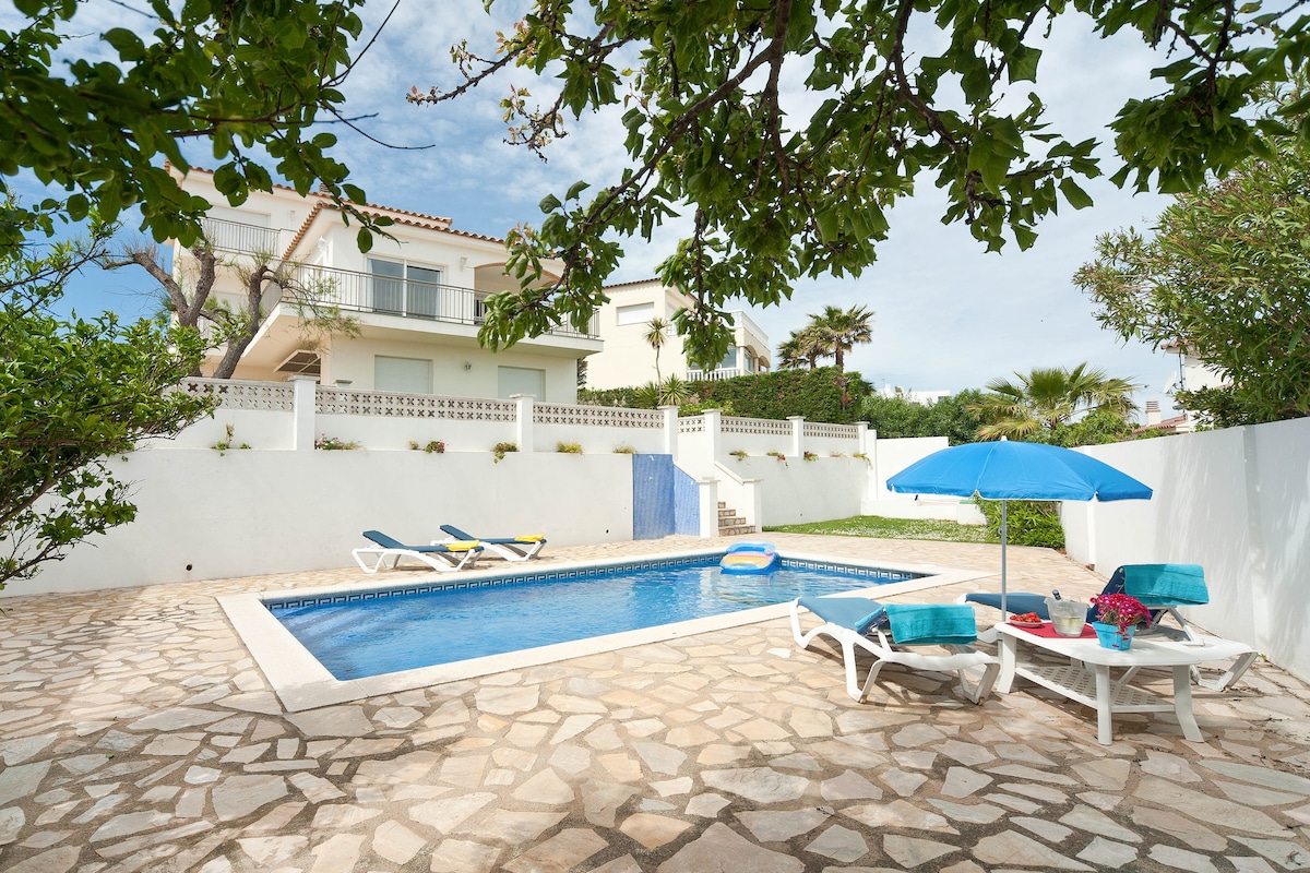 A private outdoor space features a rectangular pool, accompanied by sun loungers and a table with an umbrella. Surrounding greenery and palm trees enhance the setting, while a stone pathway and white walls provide structure to the area, ensuring a calm atmosphere for relaxation.