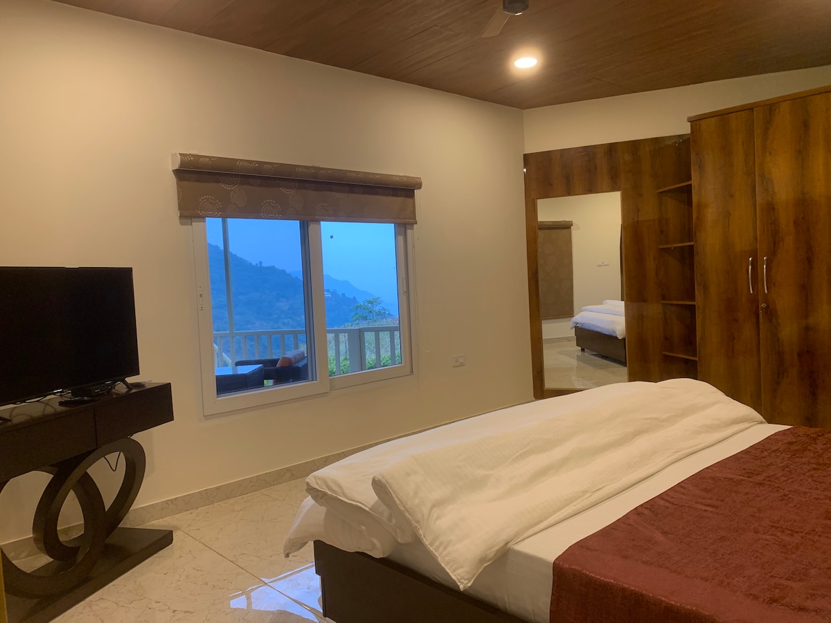 A spacious room features a comfortable bed covered with a white blanket. A television is situated on a contemporary stand. Large windows provide a view of the surrounding mountains, and a mirrored wardrobe adds depth to the space.