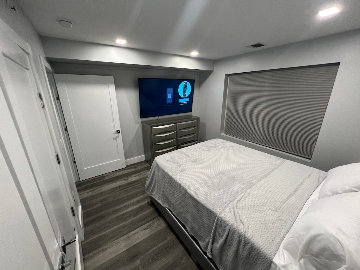 The bedroom features a spacious bed with white bedding and a large flat-screen TV mounted on the wall across from it. A dresser is positioned nearby, and natural light filters through the window, which is covered by shades. A closet door is visible on the left.