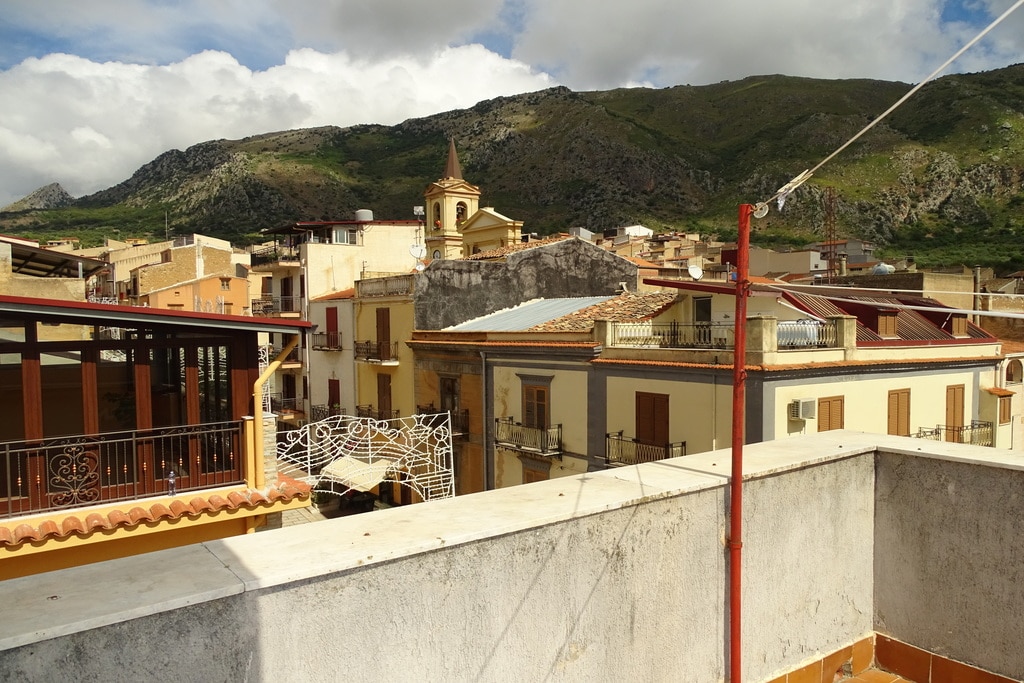 A panoramic view captures the charming rooftops of a Sicilian town, with a distant church steeple visible against a backdrop of green mountains. The scene includes a mix of traditional architecture and modern rooftops under a partly cloudy sky.