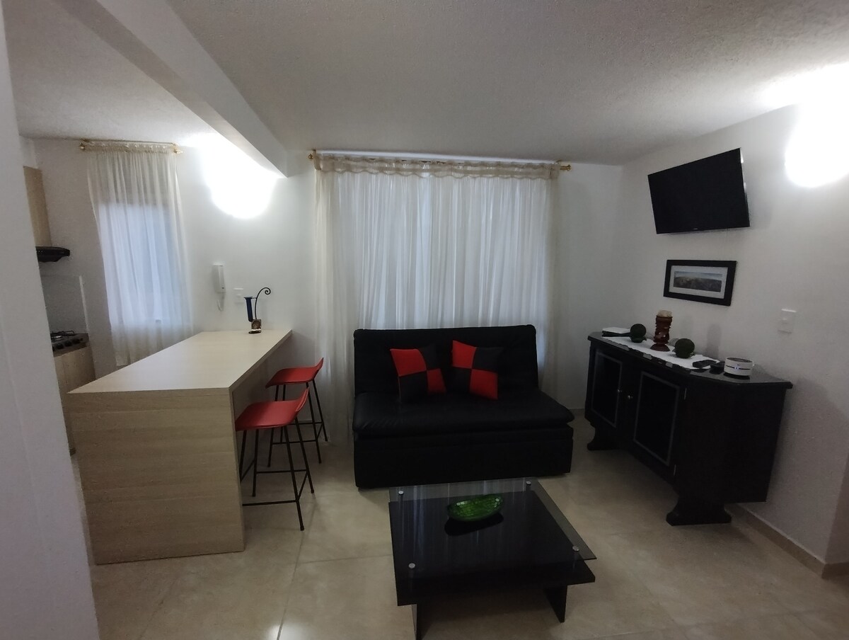 A modern living area features a black sofa adorned with red and black cushions. A glass coffee table sits in the center, with a green decorative bowl. A breakfast bar with red stools is visible, and light floods in through sheer white curtains over a window.