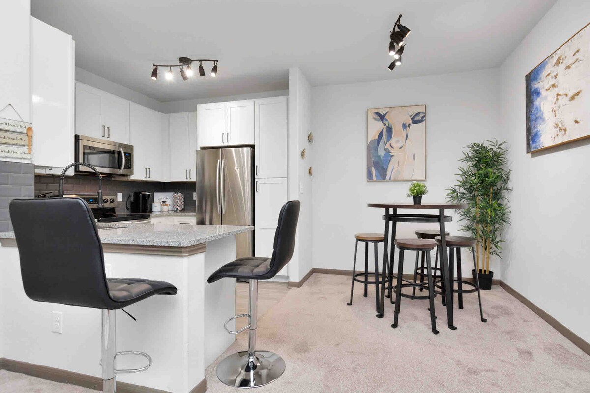 The modern kitchen area features a granite countertop with two black bar stools and stainless steel appliances. A round dining table with four wooden chairs is positioned beside a decorative plant. Light is provided by overhead track lighting, adding to the airy feel.