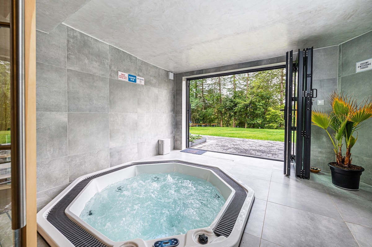 An indoor spa area features a spacious, octagonal jacuzzi with bubbling water. Large glass doors open to a serene outdoor space with green grass and trees visible. A potted plant adds a touch of nature to the corner of the room.