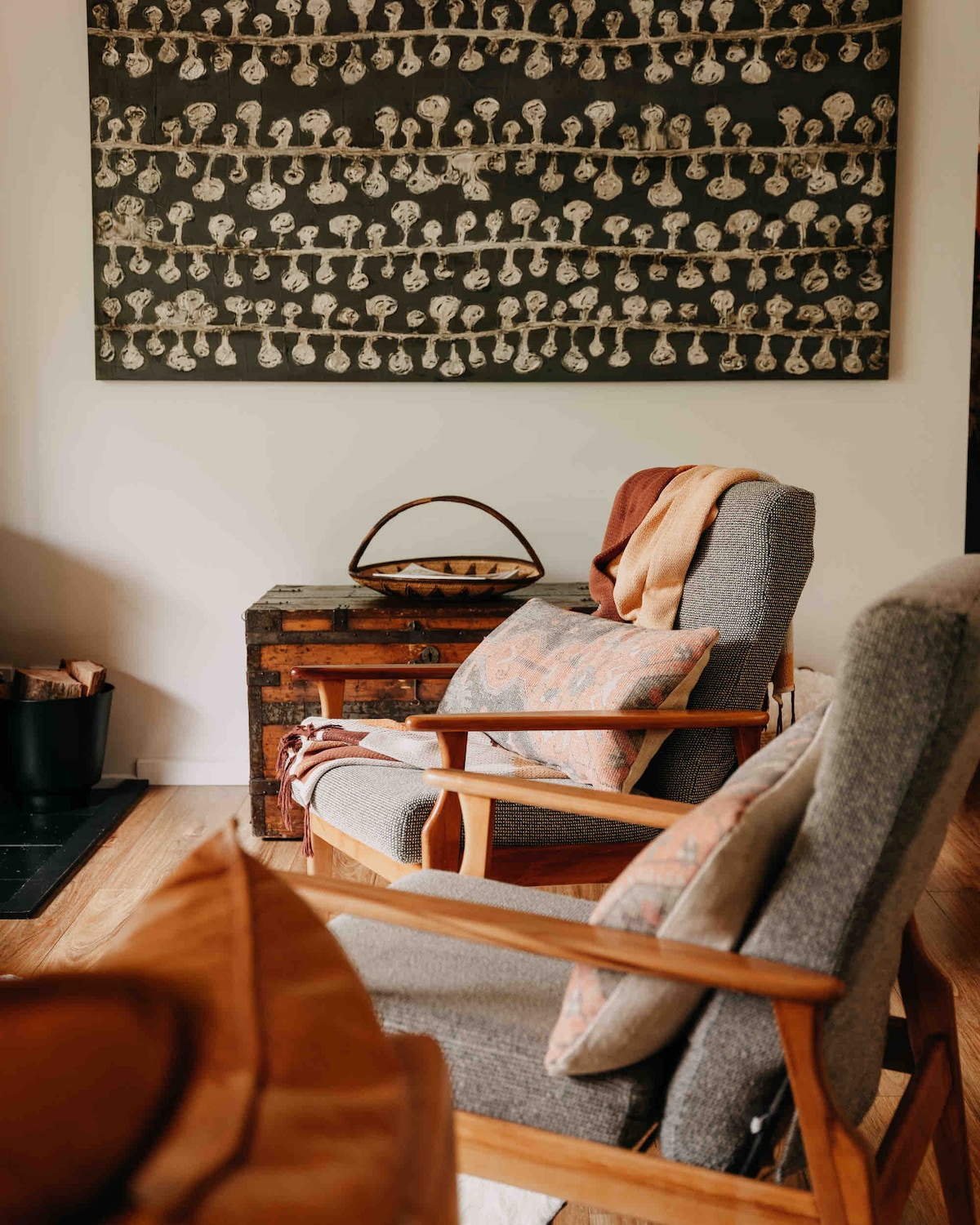 Two midcentury armchairs are positioned in a cozy corner, with decorative pillows enhancing comfort. A patterned wall hanging in neutral tones provides visual interest above a wooden side table. A small basket rests on the table, and a wood-burning fireplace is partially visible nearby.