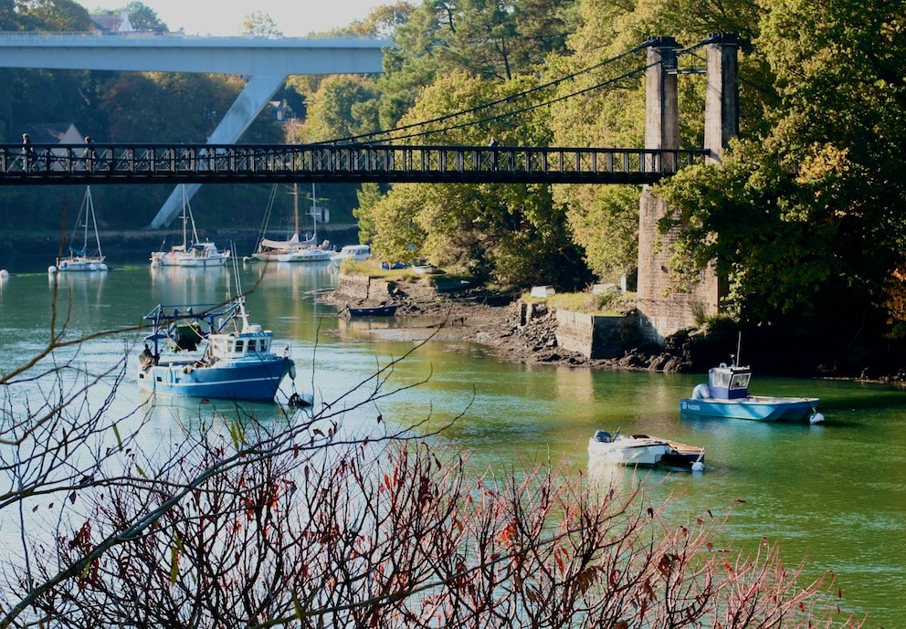 A serene waterfront scene is featured, showcasing several boats anchored in tranquil waters. A bridge spans the river in the background, framed by lush greenery. Subtle autumn colors are visible in surrounding foliage, enhancing the natural beauty of the area.