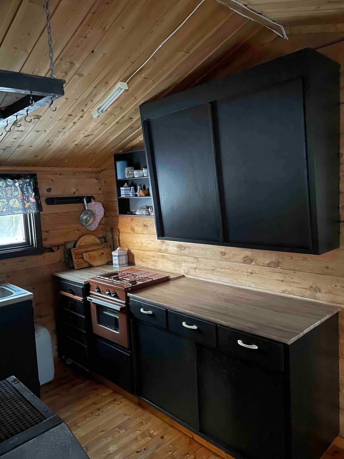 The compact kitchen features dark cabinetry and a wooden countertop. A gas stove and shelf for kitchen essentials are visible. A refrigerator is located at the far end, and a window allows natural light to enter, with a patterned curtain adding detail.