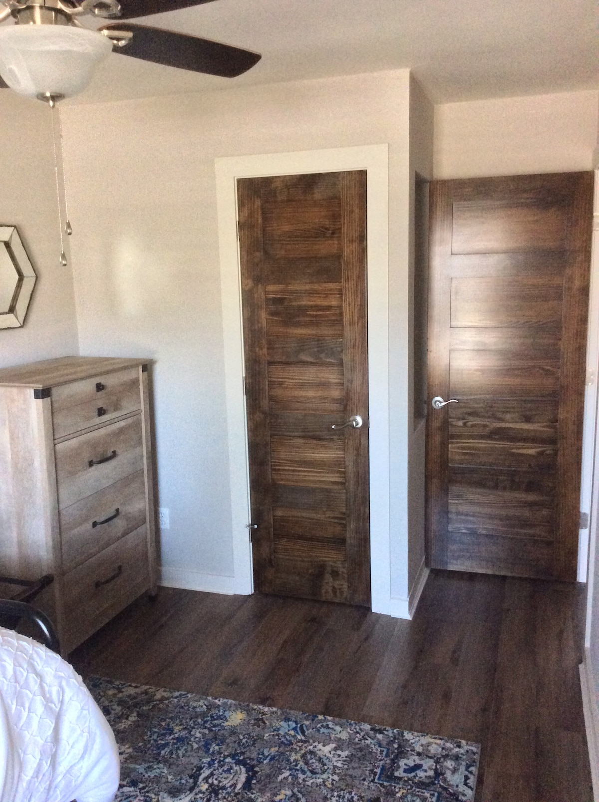 A well-lit bedroom corner is displayed, featuring a wooden dresser with five drawers. Two doors are shown, one leading to a closet and the other to a bathroom. Warm wooden textures complement the space, and stylish flooring adds to the inviting atmosphere.