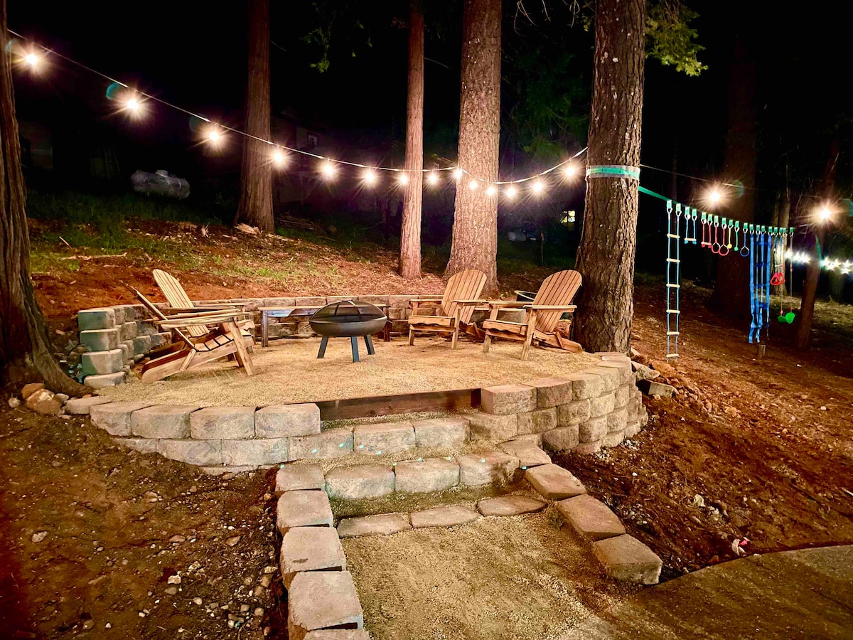 An outdoor seating area features a circular stone firepit surrounded by four wooden lounge chairs. Strung lights above create a warm ambiance in the wooded backyard, which also includes a climbing structure in the background.