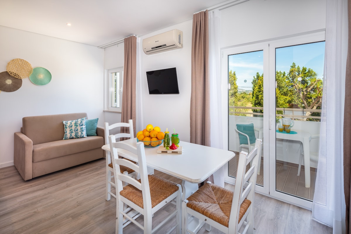 The bright living space features a sofa against the wall, paired with a white dining table surrounded by six woven chairs. A small bowl of oranges sits on the table. Large windows and a sliding door provide access to the outdoor terrace, allowing natural light to fill the room.