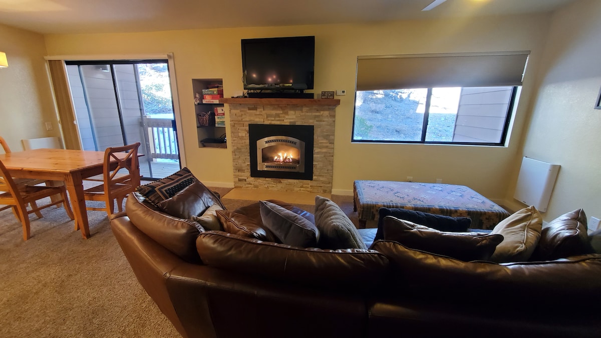 A cozy living area is presented, featuring a large leather sectional sofa adorned with decorative pillows. A stone fireplace serves as a focal point, flanked by a mounted television. Natural light enters through the windows, enhancing the warm ambience of the space.