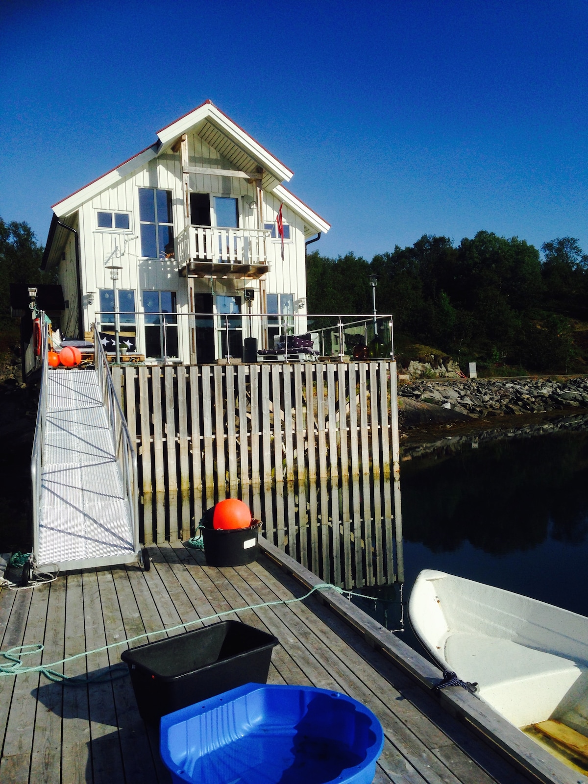 A charming rorbu stands on the quay, featuring a bright exterior and a balcony. The wooden deck leads to a gently curved ramp. A small fishing boat is anchored nearby, with fishing gear visible. The water reflects the clear blue sky, surrounded by lush greenery.