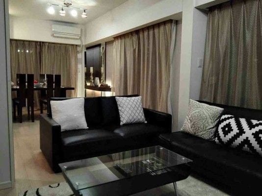 A stylish living area features two black sofas with decorative cushions, set around a glass coffee table. Soft lighting from recessed fixtures enhances the room's ambiance, while elegant curtains frame the windows, contributing to a sophisticated atmosphere.