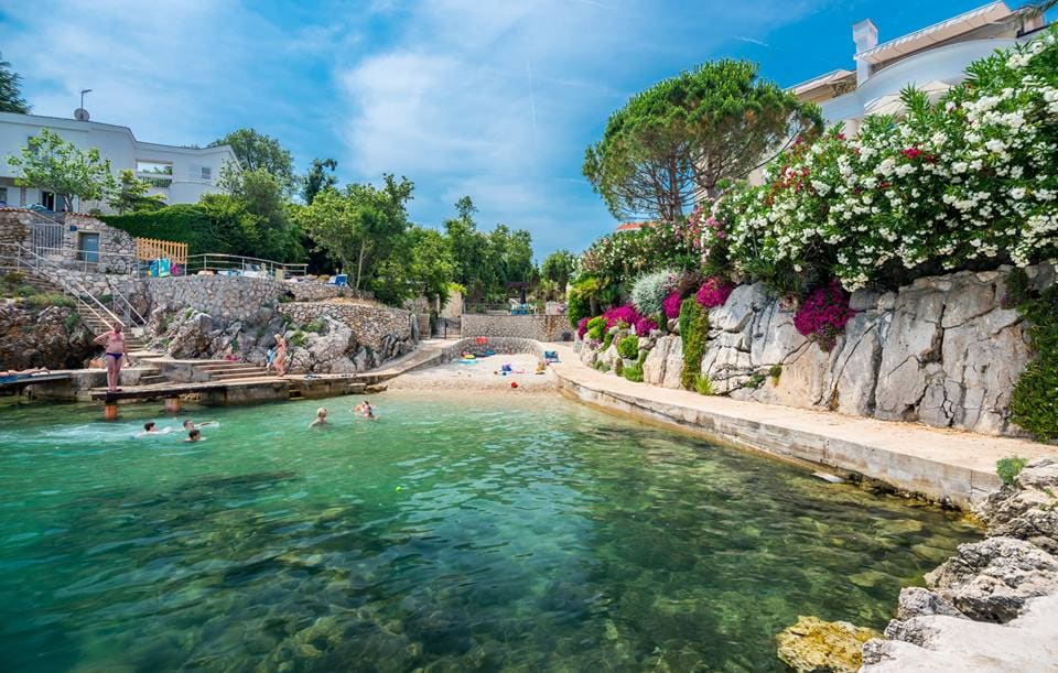 A calm, clear water cove is surrounded by lush greenery and blooming flowers. A small pebble beach is visible, along with rocky pathways leading to the shore. Several people can be seen enjoying the water, contributing to a lively atmosphere.