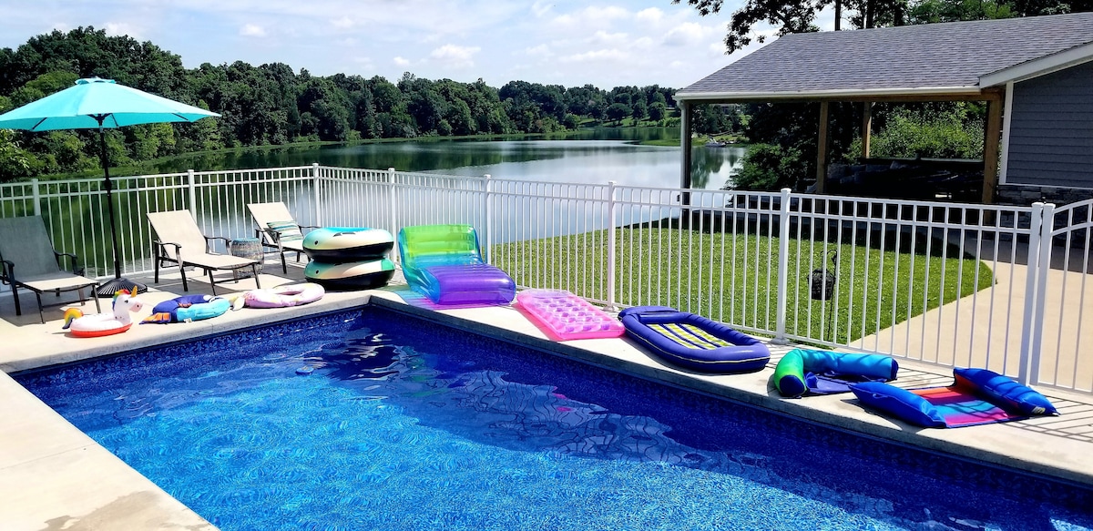 An inviting inground pool features a vibrant blue surface and is surrounded by a spacious deck. Colorful pool floats and lounge chairs are arranged near the water's edge, with a shaded umbrella providing relief from the sun. A peaceful lake view is visible in the background.