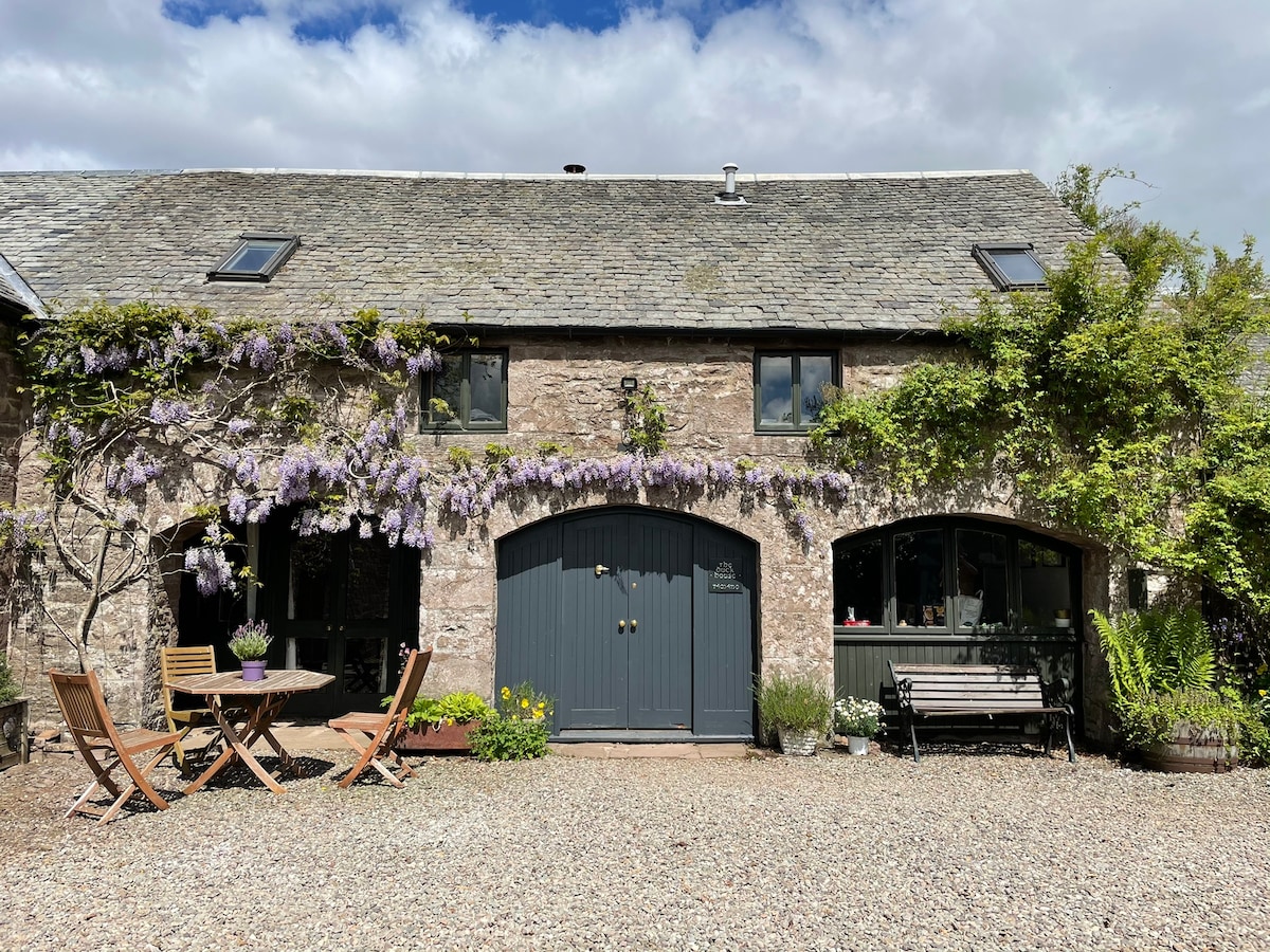 An inviting stone exterior features beautiful wisteria spilling over the entrance. A small outdoor seating area with wooden chairs and a table is positioned on a gravel courtyard, surrounded by vibrant flowers and greenery. Large windows allow natural light to enhance the welcoming atmosphere.
