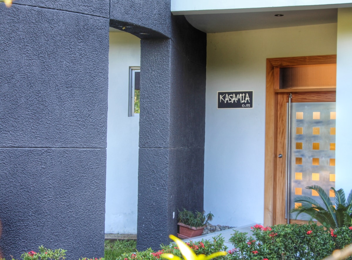 The image captures the entrance of Kasamia, featuring a modern facade with dark textured walls. A wooden door with patterned glass panels provides access, and a welcoming sign displays the name 'Kasamia' prominently. Lush greenery and colorful plants frame the entrance.
