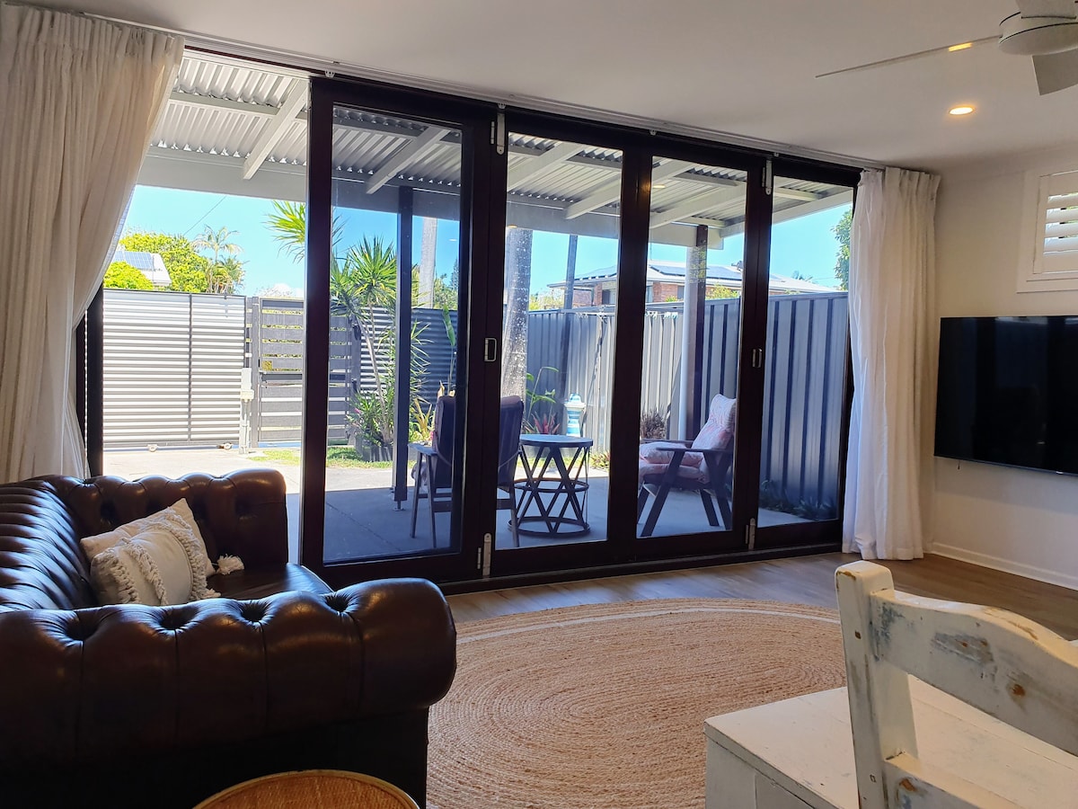 Floor-to-ceiling bi-fold doors are seen, allowing natural light to fill the bright living area. A cozy brown couch complements a round jute rug. Outside, a private patio is visible, featuring a small table and potted plants, surrounded by a fence for privacy.