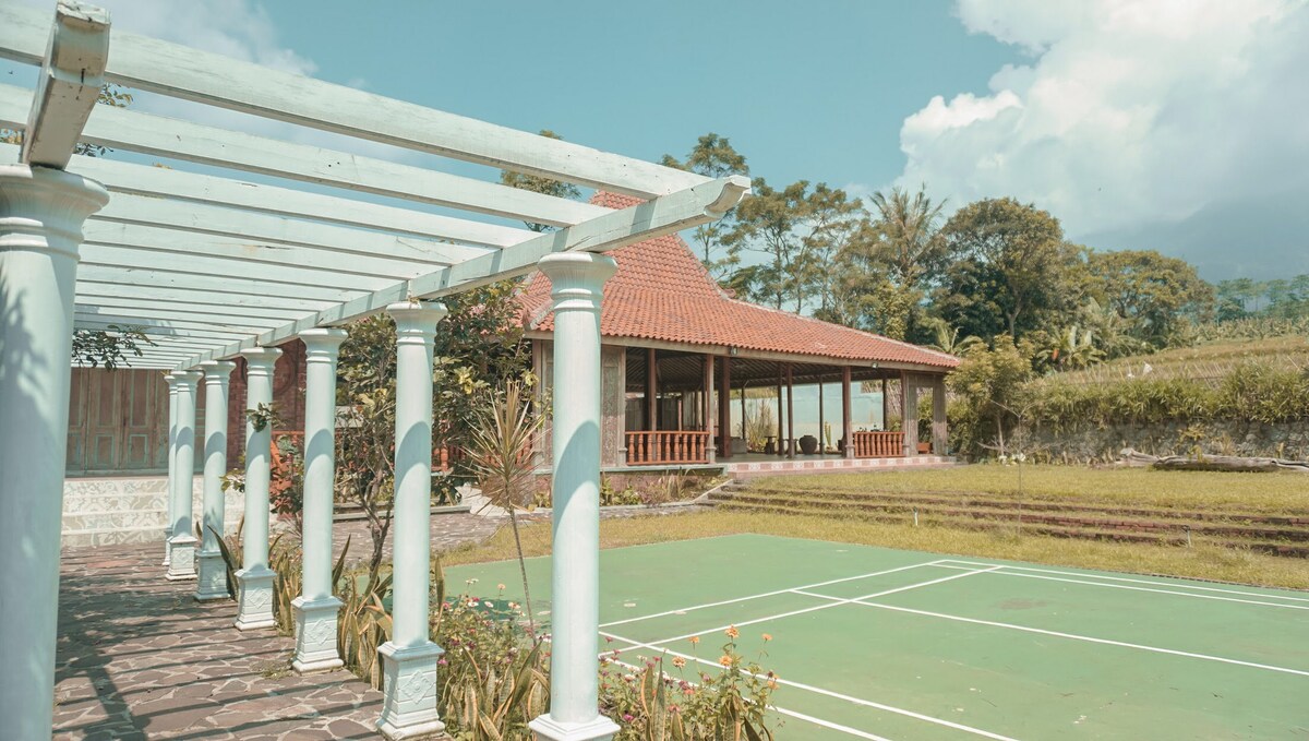 An outdoor area features a well-maintained court surrounded by greenery. A spacious pavilion with a traditional roof is visible in the background, providing shaded seating and views of the natural surroundings. Tall columns support a pergola along the pathway, enhancing the open-air ambiance.