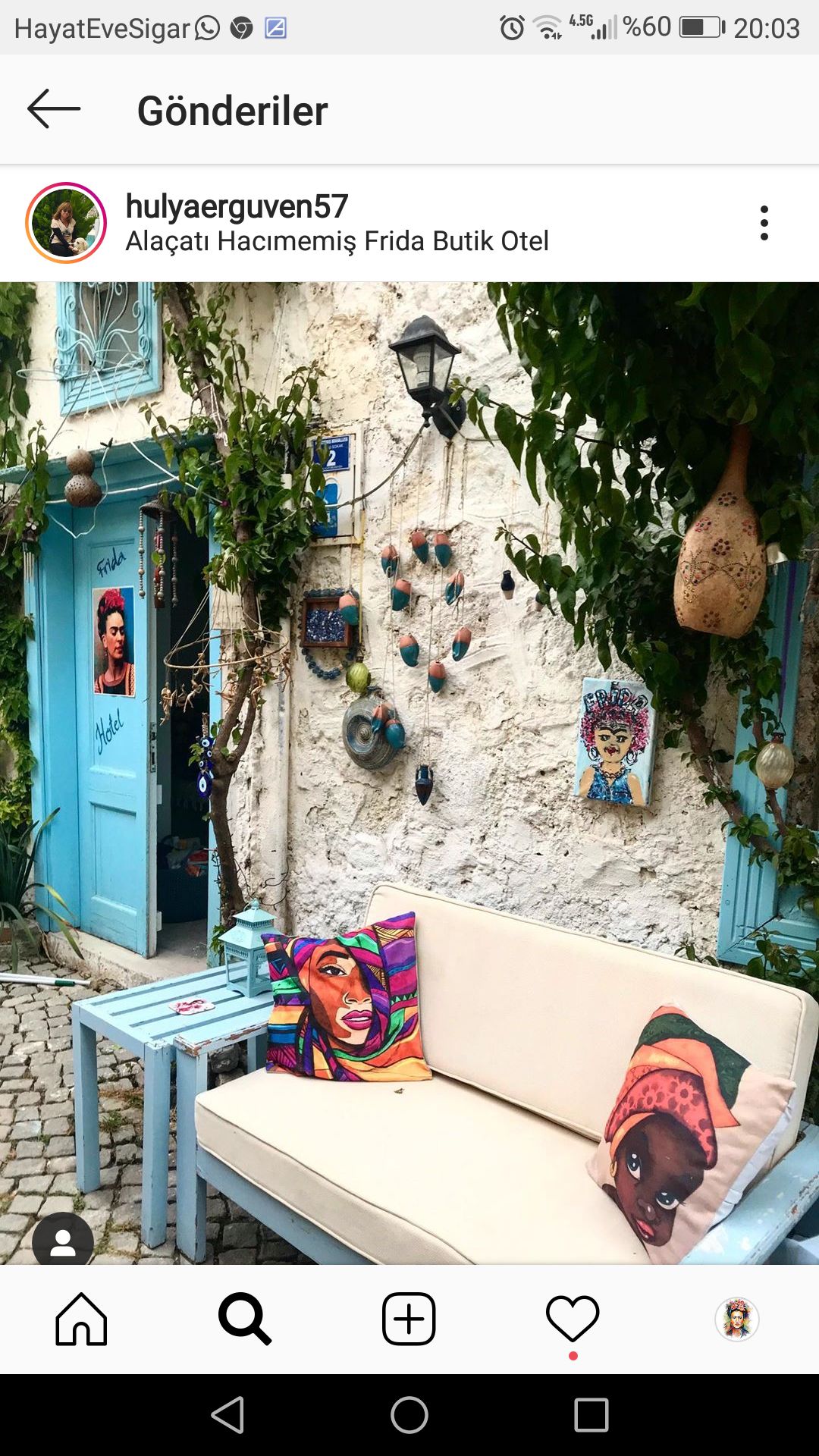 A charming outdoor seating area is set against a rustic stone wall, featuring a light-colored sofa adorned with colorful cushions. Potted plants cascade nearby, adding greenery. A vibrant blue door and various decorative elements, including painted pots, enhance the inviting atmosphere.