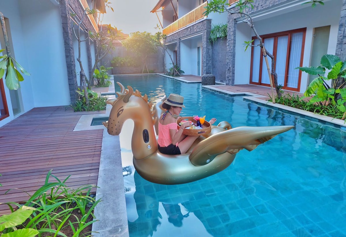 A golden inflatable float, shaped like a horse, decorates the clear waters of a swimming pool. Lush greenery and modern architecture frame the scene, while sunlight casts a warm glow over the tranquil setting, enhancing the sense of relaxation and leisure.