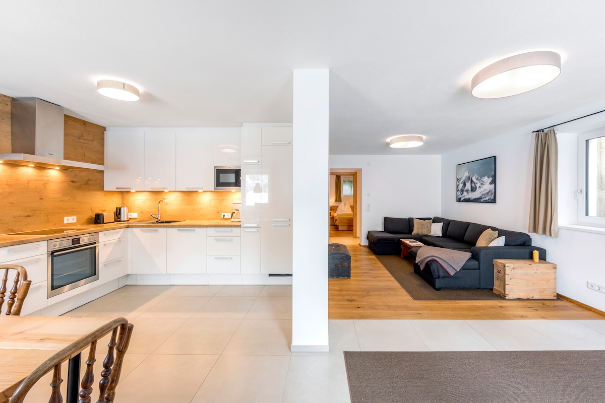 An open-concept living area features a modern kitchen with sleek cabinetry and a spacious countertop. Adjacent to the kitchen, a cozy seating area includes a dark sectional sofa and a wooden coffee table. Natural light filters through the nearby window, creating a bright and welcoming space.