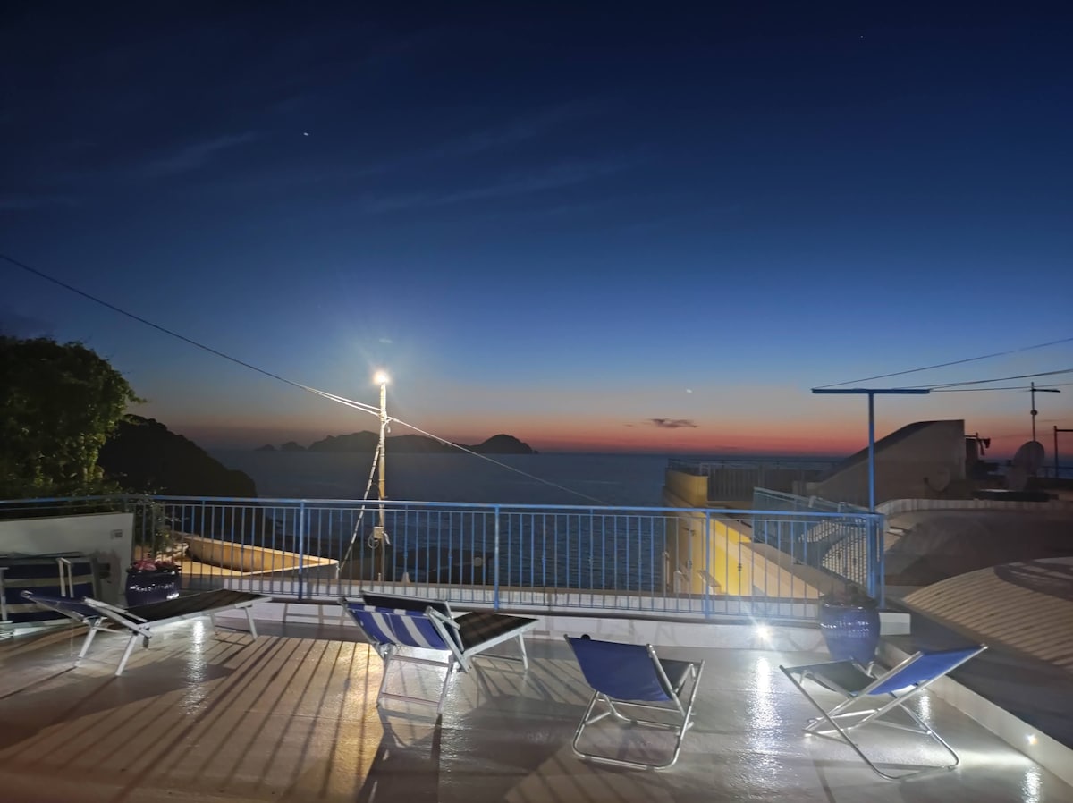 An expansive terrace is illuminated under the night sky, featuring several lounge chairs. The view encompasses the sea and distant islands, with a gradient of colors in the horizon from deep blue to hints of orange. Soft lighting adds a serene ambiance.