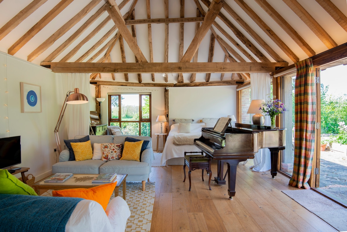 The open-plan living space features oak beams overhead, with a comfortable sitting area including a sofa and armchair. A grand piano is positioned nearby, and large glass doors open to the terrace, allowing natural light to fill the room. A cozy sleeping area is discreetly arranged behind a curtain.