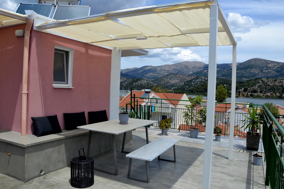 A rooftop terrace features a shaded seating area with a table and black cushions. Surrounding views include hills and nearby rooftops. The terrace is outfitted with potted plants and a black lantern, providing a tranquil space for relaxation.