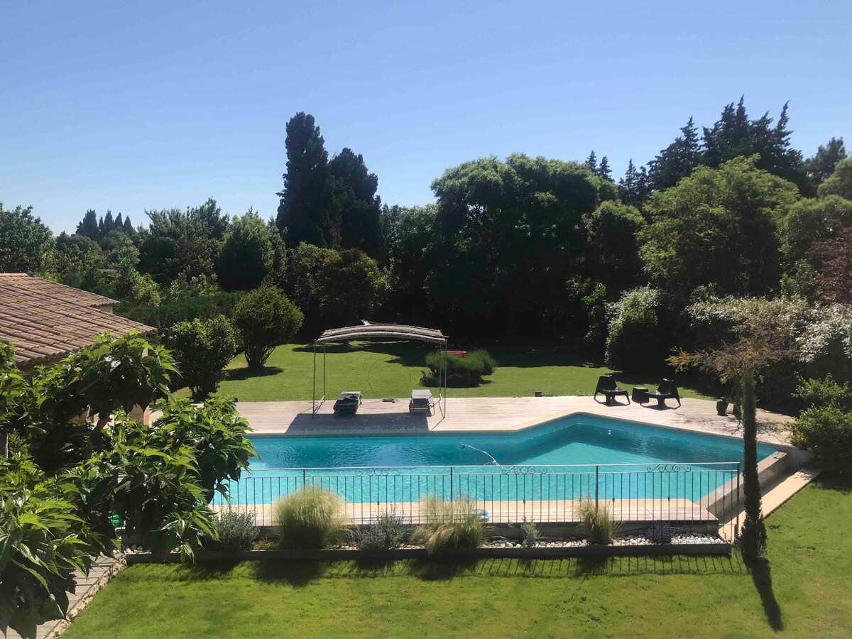 A well-maintained outdoor area features a large swimming pool surrounded by lush greenery. Comfortable lounge chairs are positioned near the water, while a shaded pergola provides a relaxing seating option. Trees and shrubs create a private and serene atmosphere.