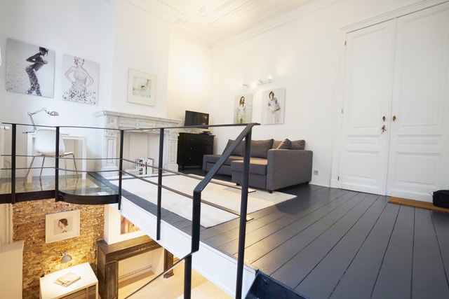 A bright and open living area features a gray sofa facing a wall-mounted television. A small desk is positioned against one wall. The space is divided by a staircase leading to a mezzanine level, showcasing stylish artwork and a combination of light and dark flooring.