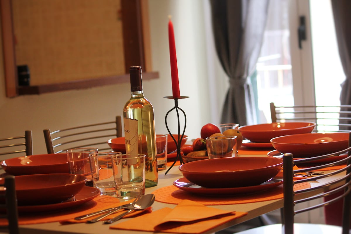 A dining area is set with an arrangement of orange plates and glasses on a white table. A single red candle is placed on a holder in the center, accompanied by a small bowl of fruits. Natural light filters through nearby curtains.