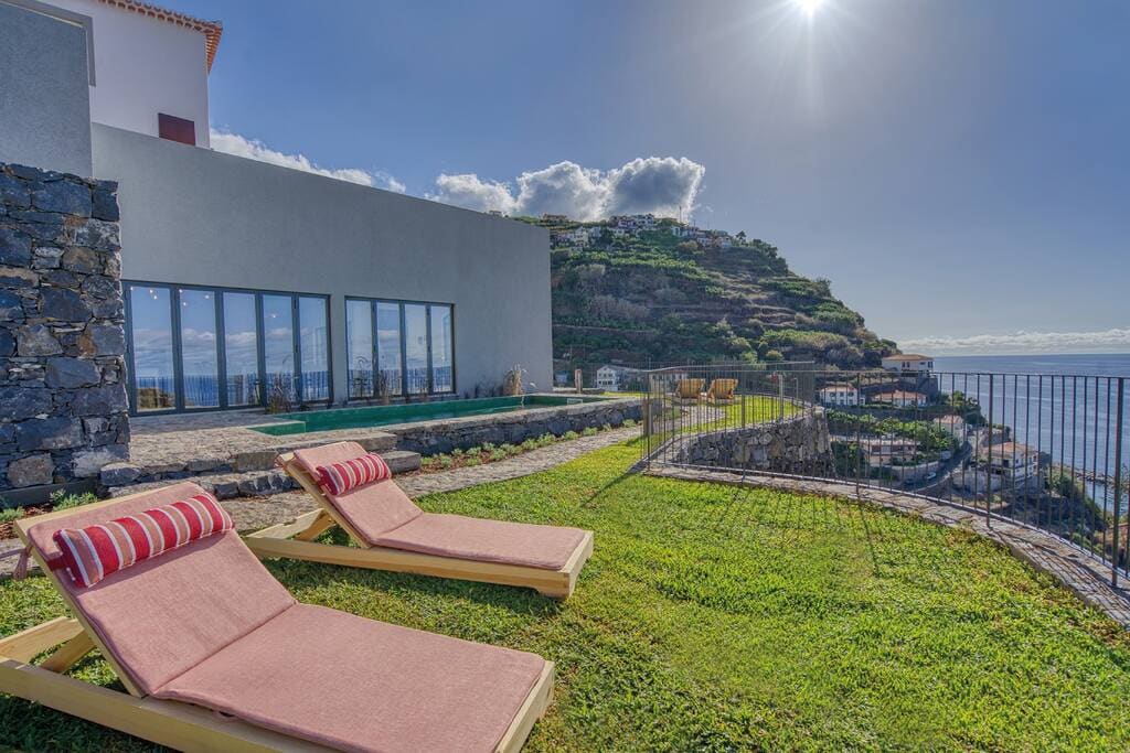 Sun loungers with striped cushions are positioned on green grass, facing expansive glass doors that open to a contemporary building. The horizon displays a scenic view of the sea and terraced hills, illuminated by bright sunlight.