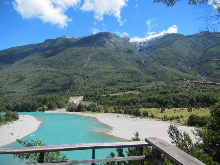 A serene landscape is captured, featuring a vibrant turquoise river winding through a valley surrounded by lush greenery. Majestic mountains rise in the background, their peaks partially covered with snow under a clear blue sky dotted with clouds.