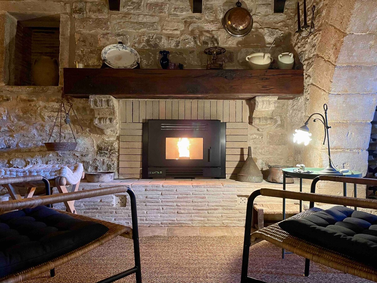 A cozy living area features a rustic stone fireplace at its center, surrounded by a wooden mantel adorned with culinary and decorative items. Two wicker chairs with soft cushions invite relaxation, while a warm glow emanates from the fire, creating an inviting atmosphere.