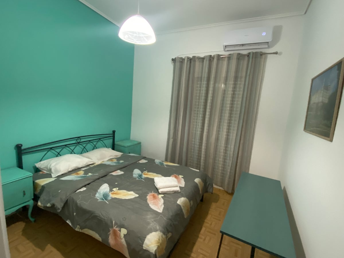 A bedroom is presented with vibrant green walls and a spacious bed dressed in a floral patterned cover. Two side tables flank the bed, and a wooden desk is positioned along one wall. Sheer curtains allow soft light to filter into the room.