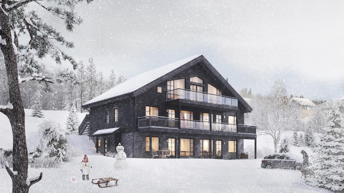 The exterior of a modern chalet is shown, surrounded by snow-covered grounds. A spacious balcony with multiple levels overlooks the winter landscape. Snowflakes gently fall, while a snowman and sled are visible in the foreground, enhancing the seasonal charm of the property.