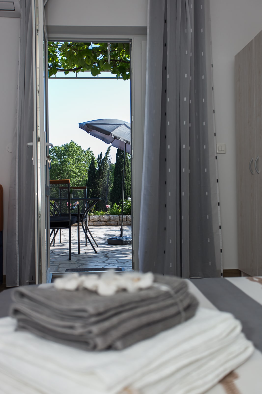 The image showcases a view from inside the apartment looking out towards a common terrace. A set of outdoor furniture, including a table and chairs, is visible under a large umbrella. Soft curtains frame the doorway, enhancing the inviting atmosphere.