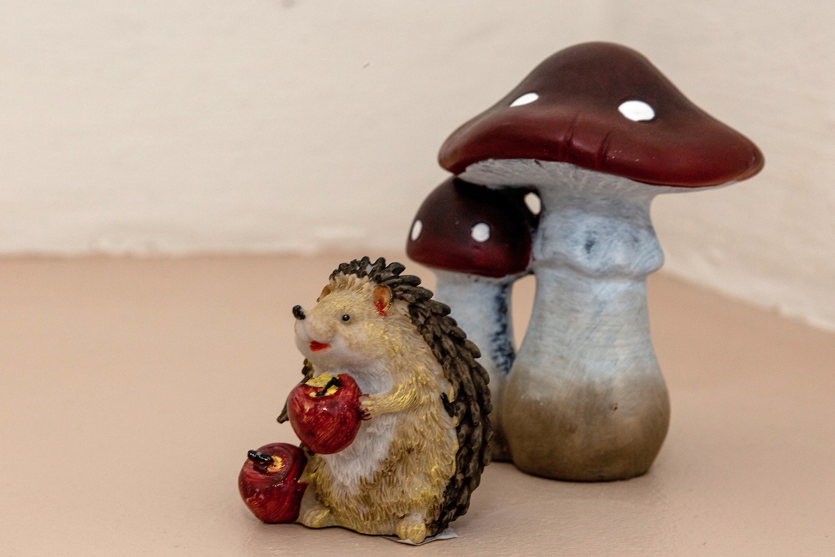A decorative arrangement features a small hedgehog figurine holding two red apples, positioned beside two stylized mushrooms. The mushrooms, with maroon caps and white spots, create a whimsical contrast against the smooth, neutral-colored surface.