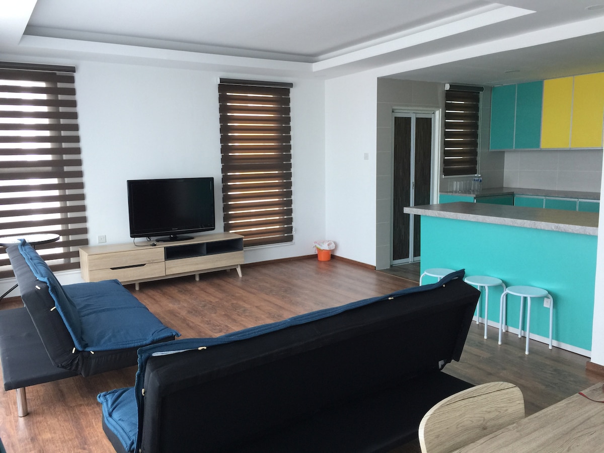 A bright living area showcases a modern design with large windows featuring brown striped blinds. Two dark couches are positioned opposite a television on a wooden console. A colorful kitchen area is visible in the background, highlighted by shades of turquoise and yellow cabinetry.